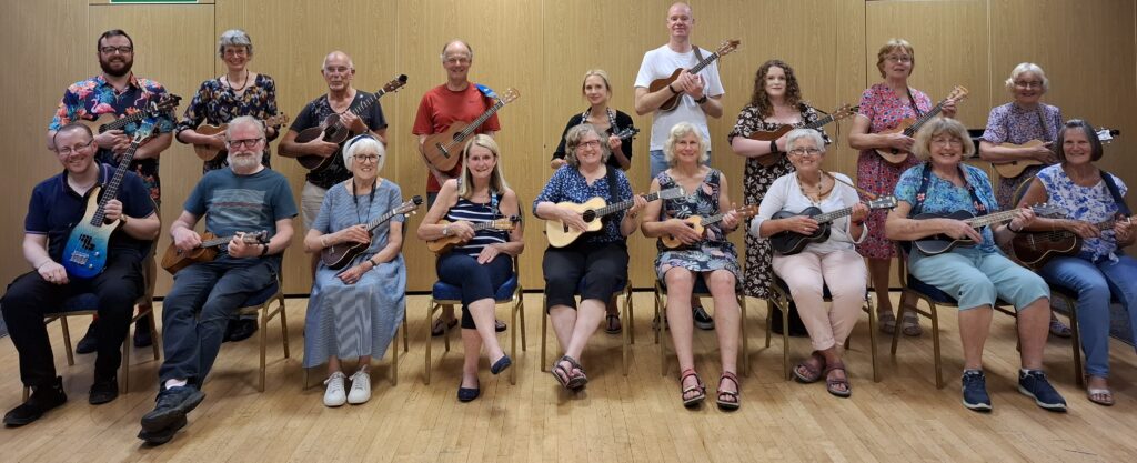 A group of musicians in two rows. The front row is seated, the rear row is standing. They are all holding ukuleles.