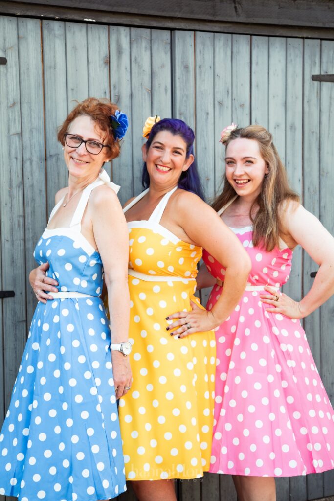 Three females singers in bright coloured polka dot dresses. From the left, the dresses are blue with white spots, yellow with wite spots and pink with white spots.