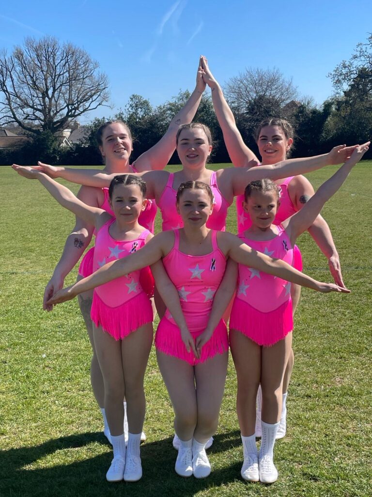 A group of Majorettes in pink uniforms. The Majorettes are stanging in a close group and using their arms to make a star shape.